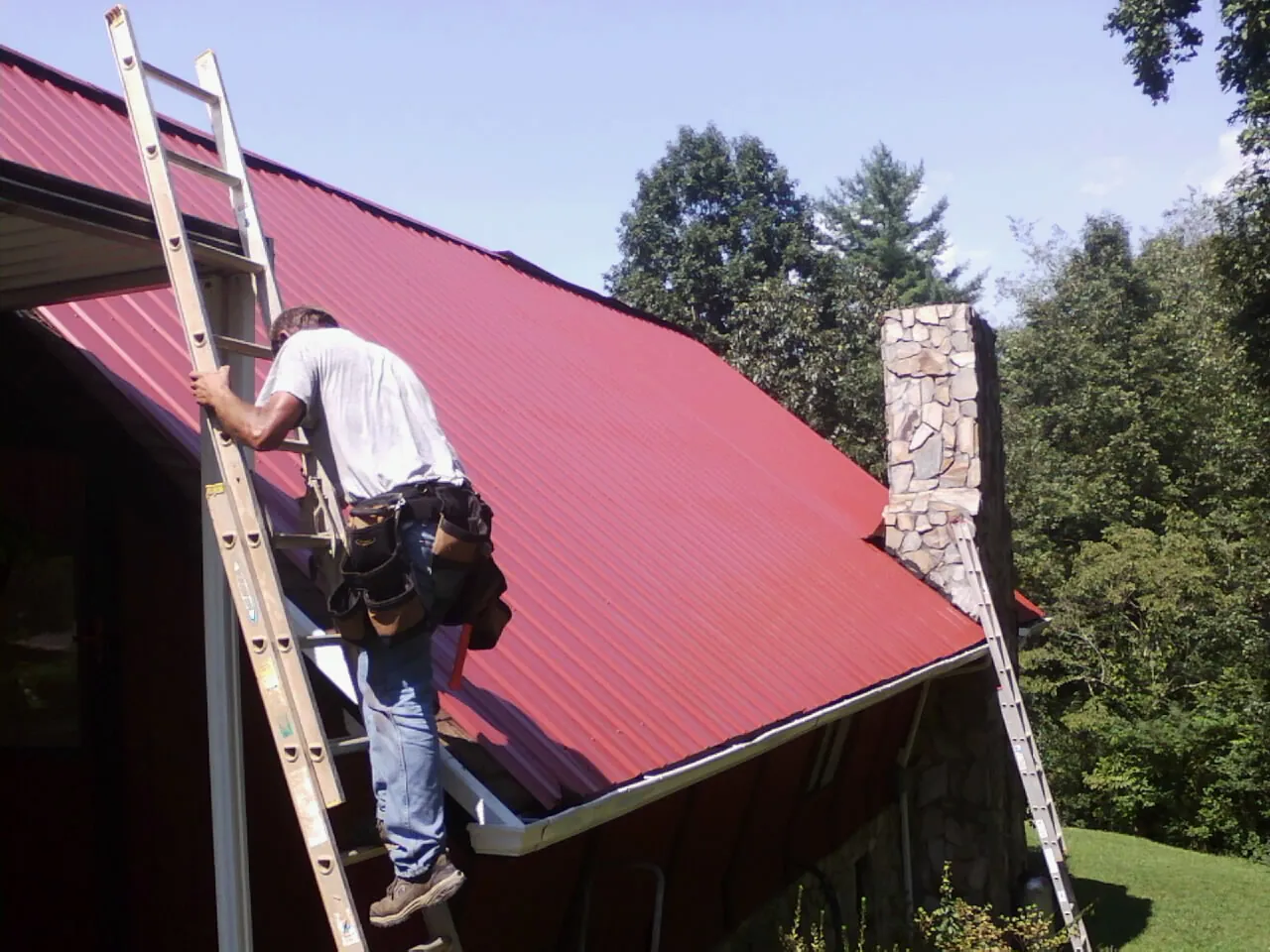 Roofer on a ladder beside a red metal roof and stone chimney — Soffit & Fascia Repair by Roofing Brothers in Smelterville